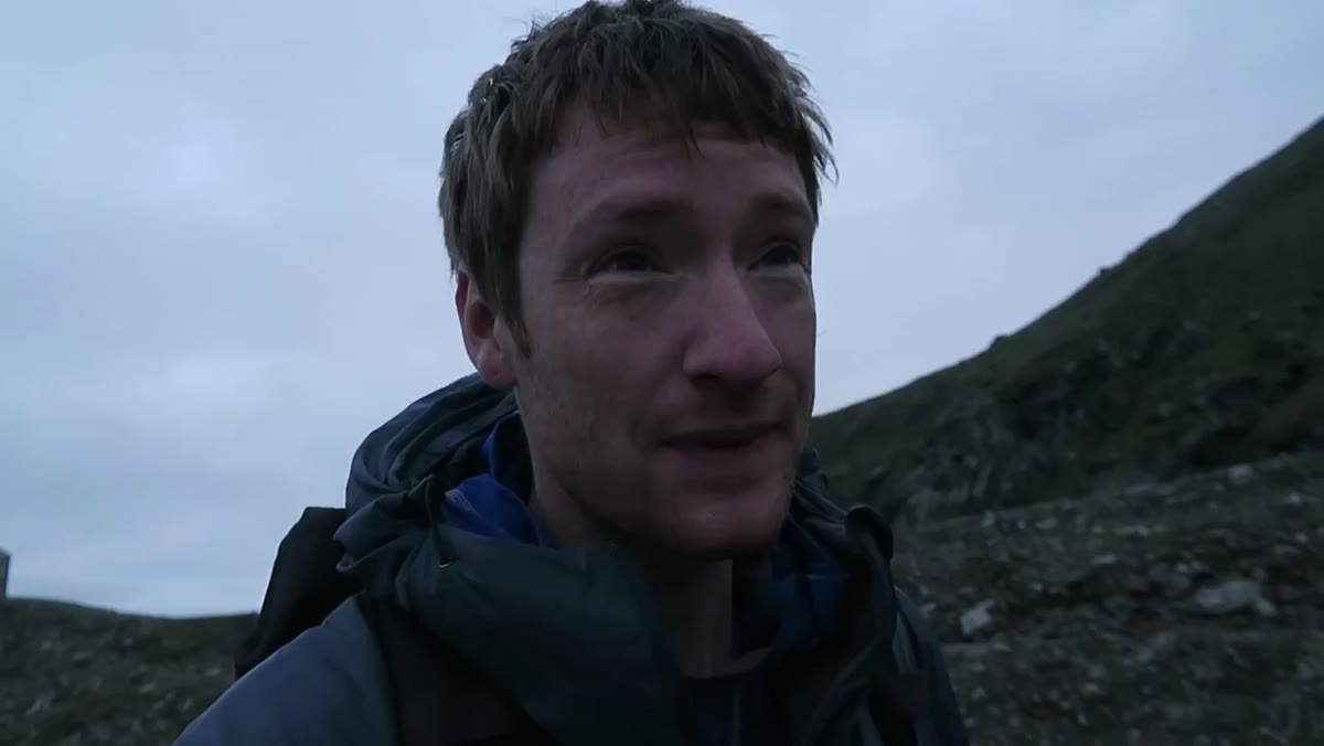 Heaton speaking outside hut, mist clearing behind him
