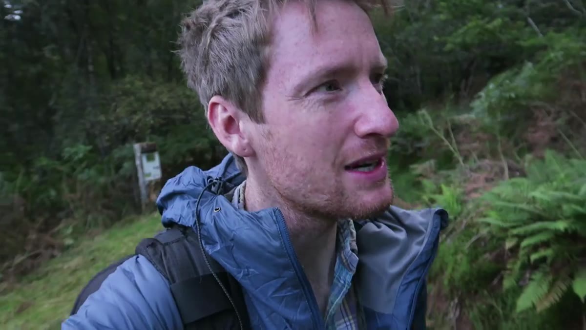 Heaton moving through woodland, scanning surroundings, camera at his side