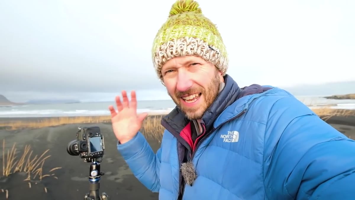 Danson crouching near beach grasses with black sand and cliffs behind
